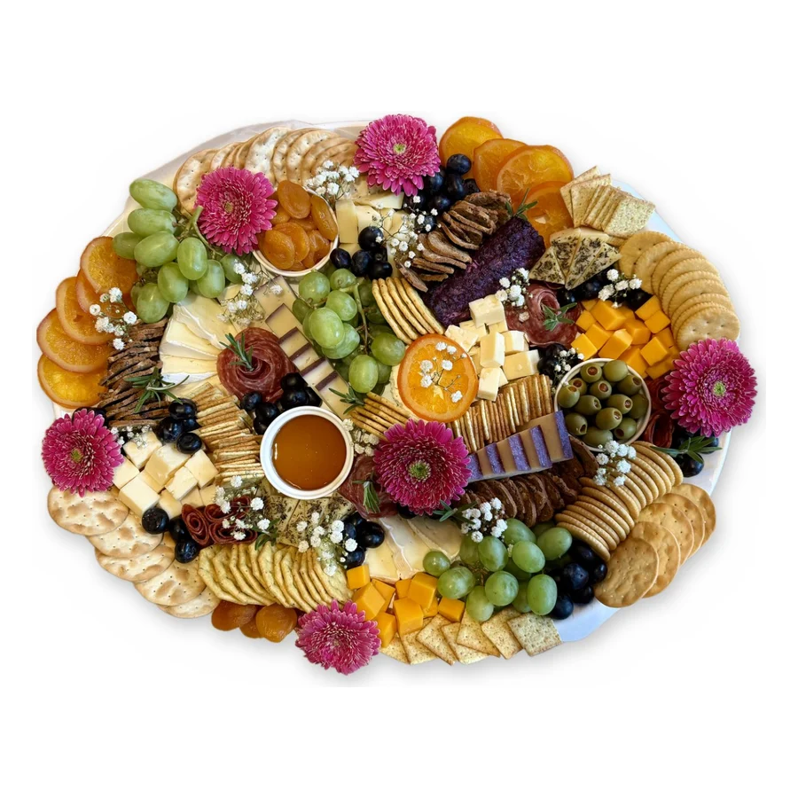 A large oval charcuterie board with pink gerbera daisies, cheeses, salami roses, grapes, candied oranges, crackers and a bowl of honey